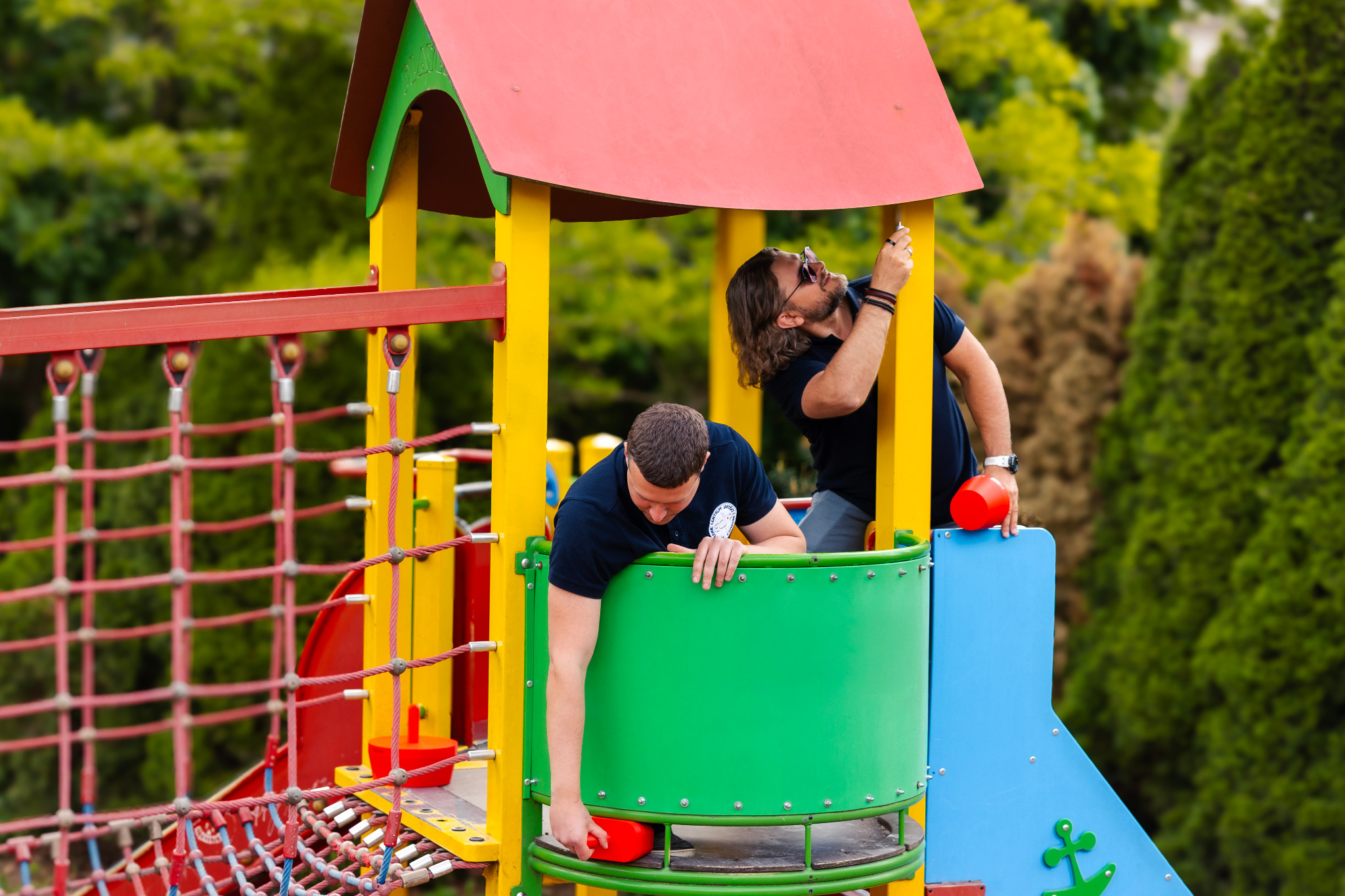 Dwóch mężczyzn w trakcie inspekcji placu zabaw z czerwonymi elementami. Jeden sprawdza konstrukcję dachu, drugi pochyla się nad zieloną platformą.