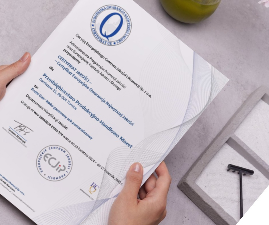 Osoba trzyma certyfikat jakości 'Europejska Gwarancja Najwyższej Jakości' dla firmy Maxet, na tle dekoracyjnego ogrodu zen i napoju.