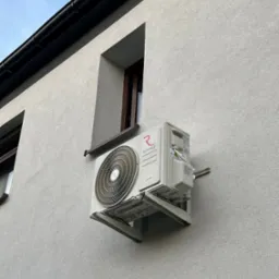 Biała klimatyzacja Rotenso zamontowana na szarej elewacji budynku, widoczna z boku, z fragmentami okien i dachu. Kąt ujęcia od dołu.