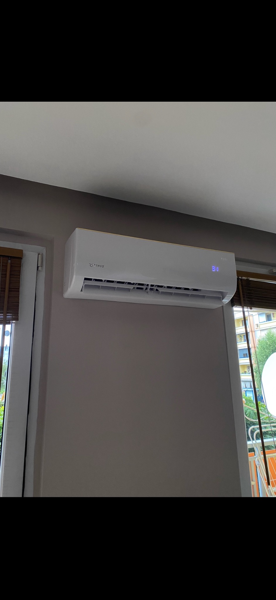 Biały klimatyzator marki Tensa zamontowany na szarej ścianie, widoczny fragment okna z drewnianymi żaluzjami i balkon za nim.