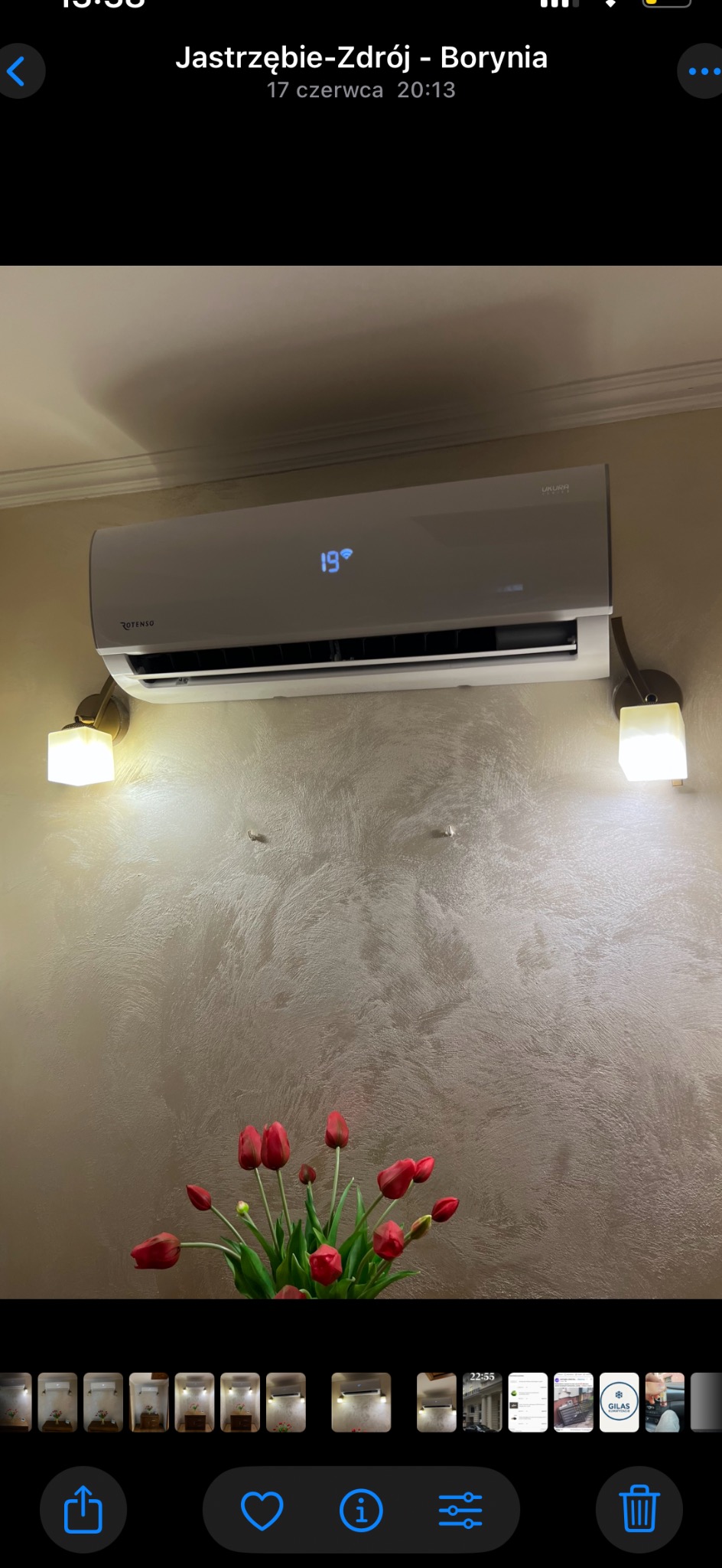 Biały klimatyzator Rotenso Ukura z wyświetlaną temperaturą 19 stopni Celsjusza, zamontowany na ścianie z dekoracyjnym tynkiem, oświetlony lampkami i ozdobiony bukietem czerwonych tulipanów.