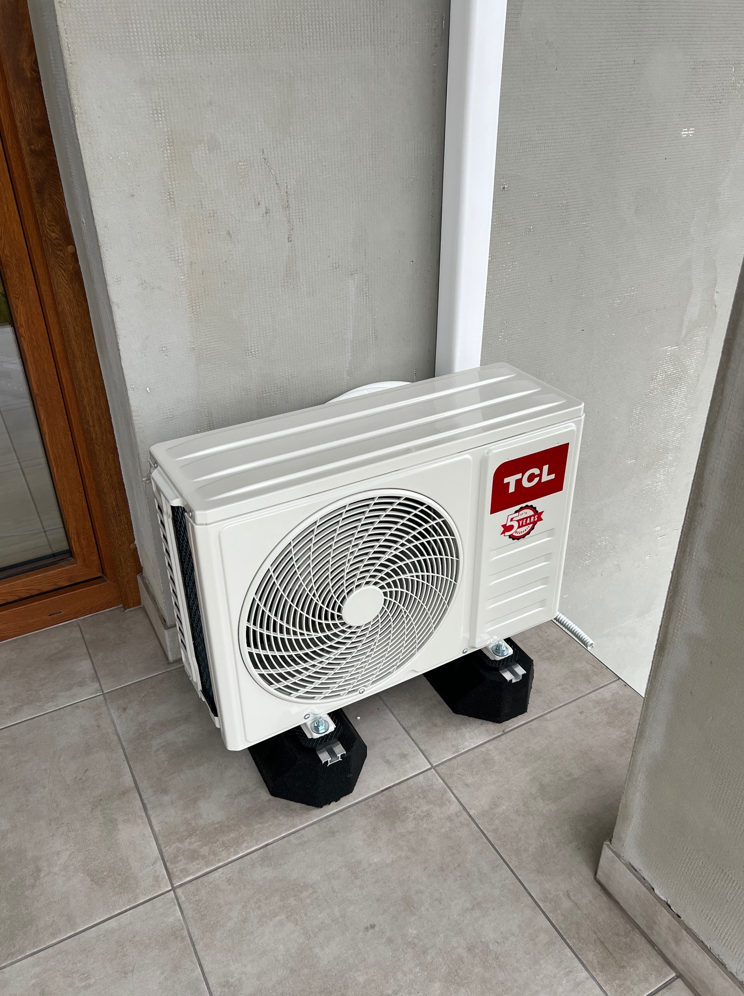 Biała jednostka zewnętrzna klimatyzacji TCL zamontowana na czarnych podkładkach antywibracyjnych, z widocznym przewodem odprowadzającym skropliny.