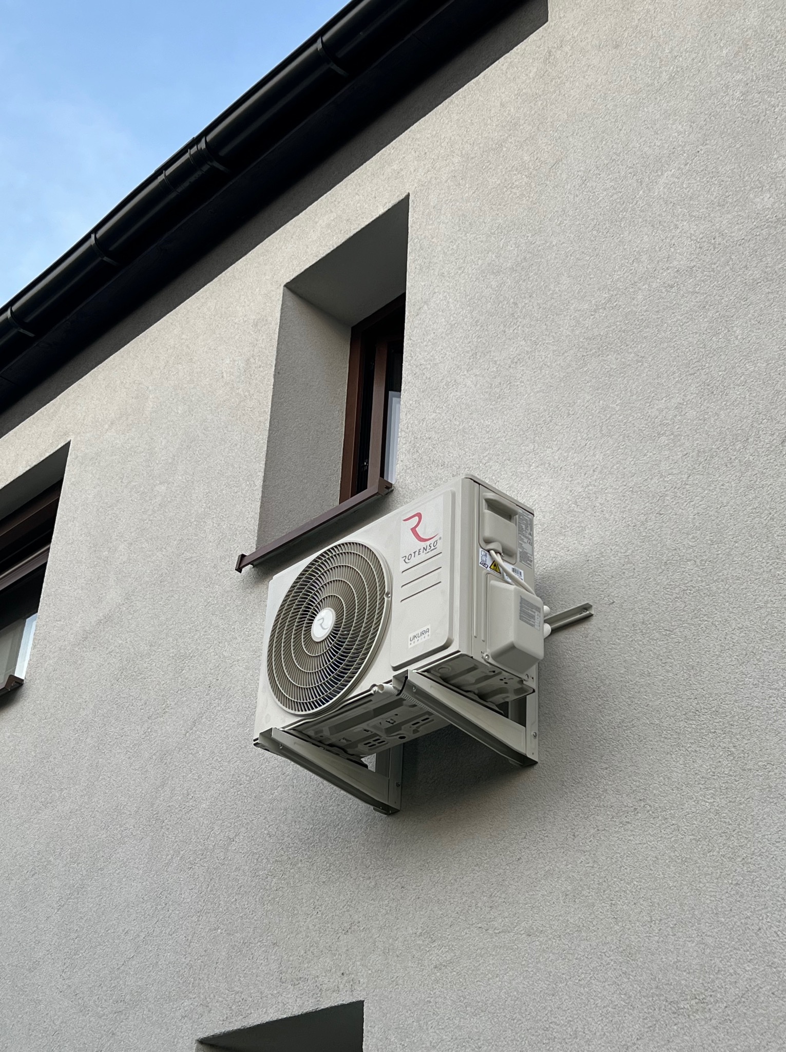 Biała klimatyzacja Rotenso zamontowana na szarej elewacji budynku, widoczna z boku, z fragmentami okien i dachu. Kąt ujęcia od dołu.