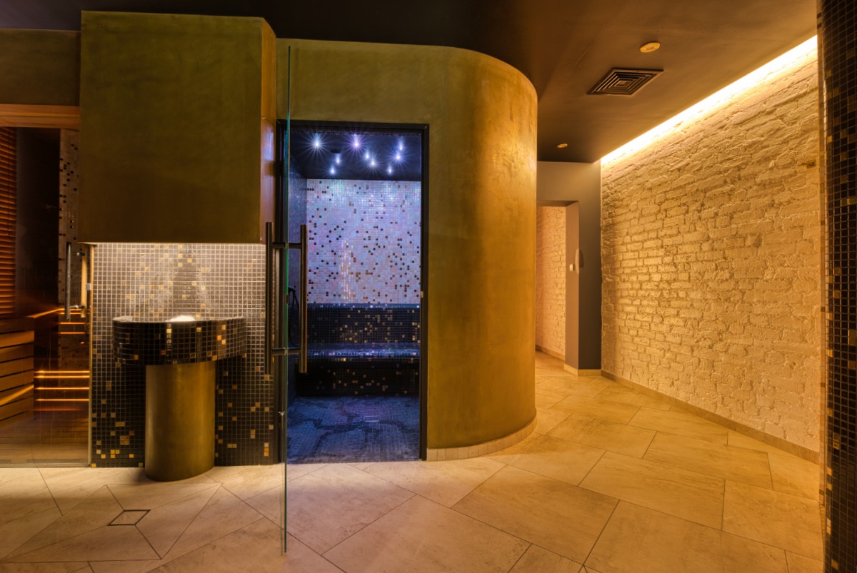 Nowoczesne wnętrze spa z mozaikami, oświetleniem LED i elementami z naturalnego kamienia. Widoczna sauna, umywalka i ściana z cegły. Ciepłe oświetlenie.