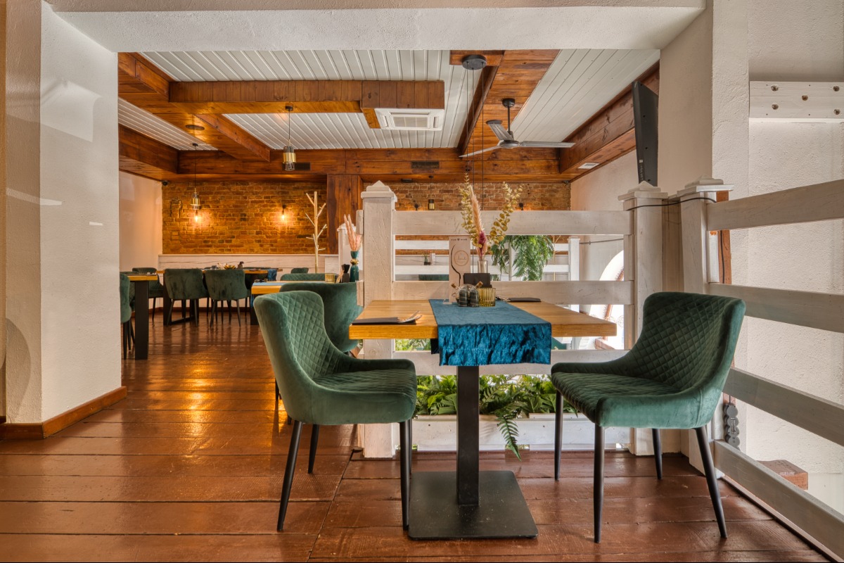 Przytulne wnętrze restauracji: drewniane belki sufitowe, ceglana ściana, zielone krzesła i nakryty stół. Ciepłe światło podkreśla detale wystroju.