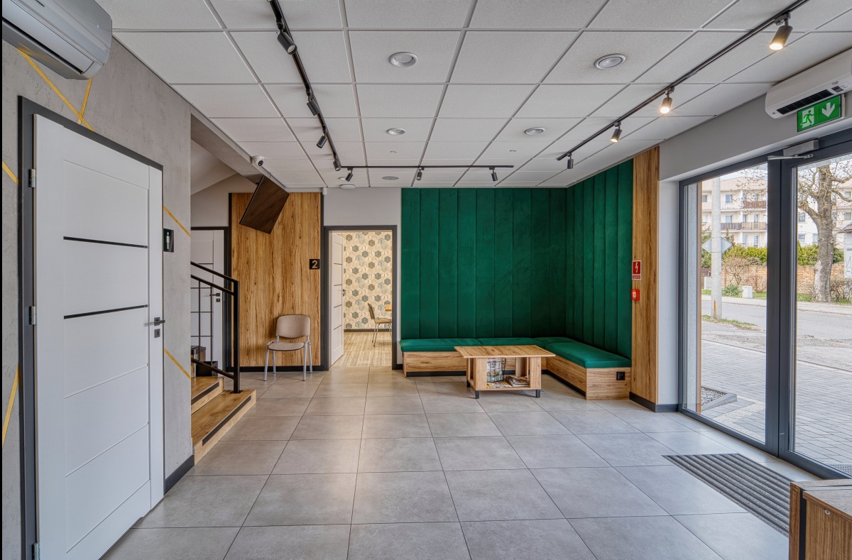 Nowoczesne wnętrze biurowe z zieloną ścianą, drewnianymi akcentami i szarą podłogą. Widoczne drzwi, schody i fragment ulicy za oknem.