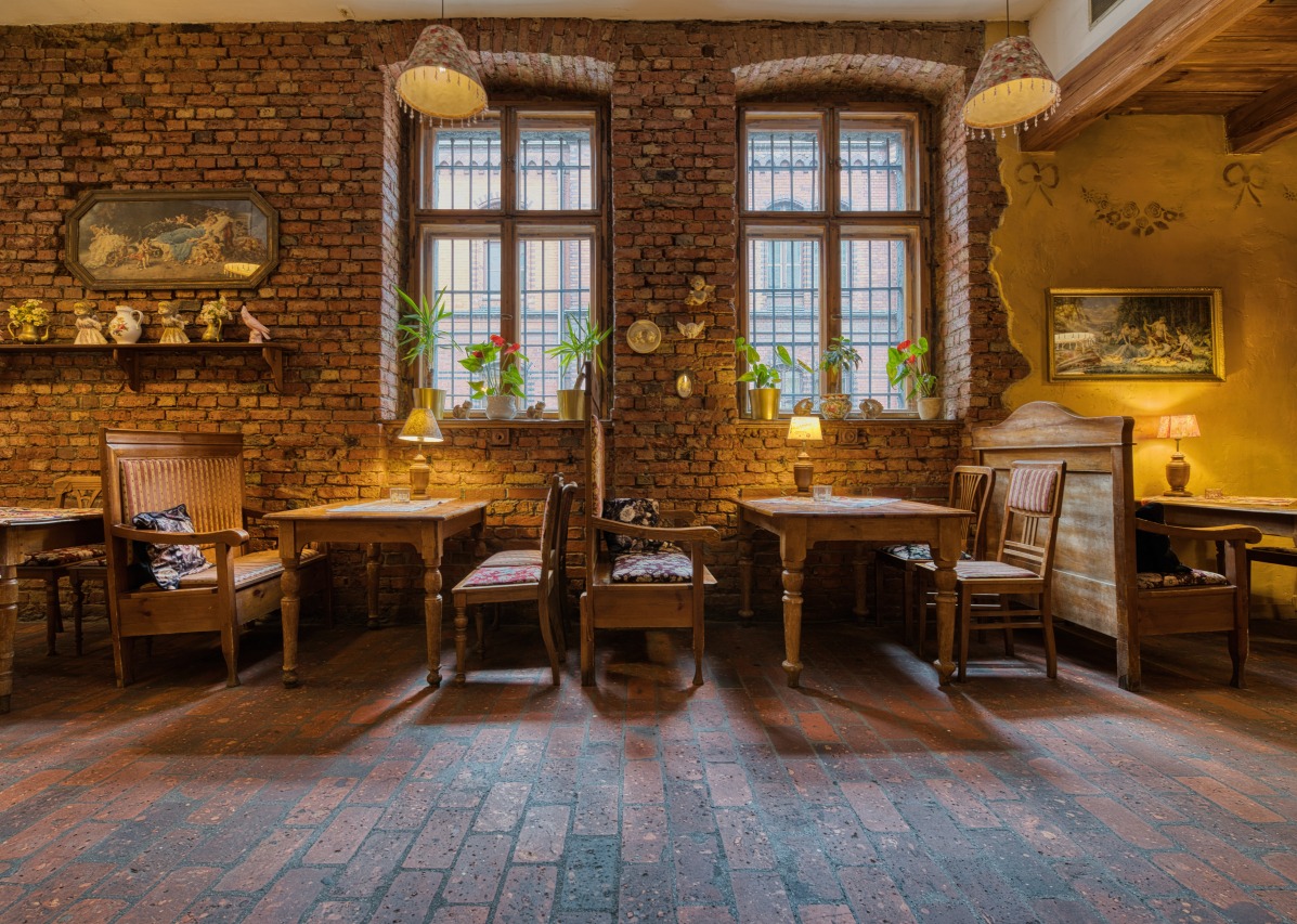 Rustykalne wnętrze restauracji z ceglanymi ścianami, drewnianymi stołami i krzesłami, oświetlone lampami stołowymi i wiszącymi. Klimatyczny wystrój z obrazami i roślinami.
