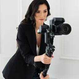 Kobieta w czarnym żakiecie trzyma stabilizator z aparatem, przygotowując się do nagrania. Jasne tło, profesjonalny sprzęt.