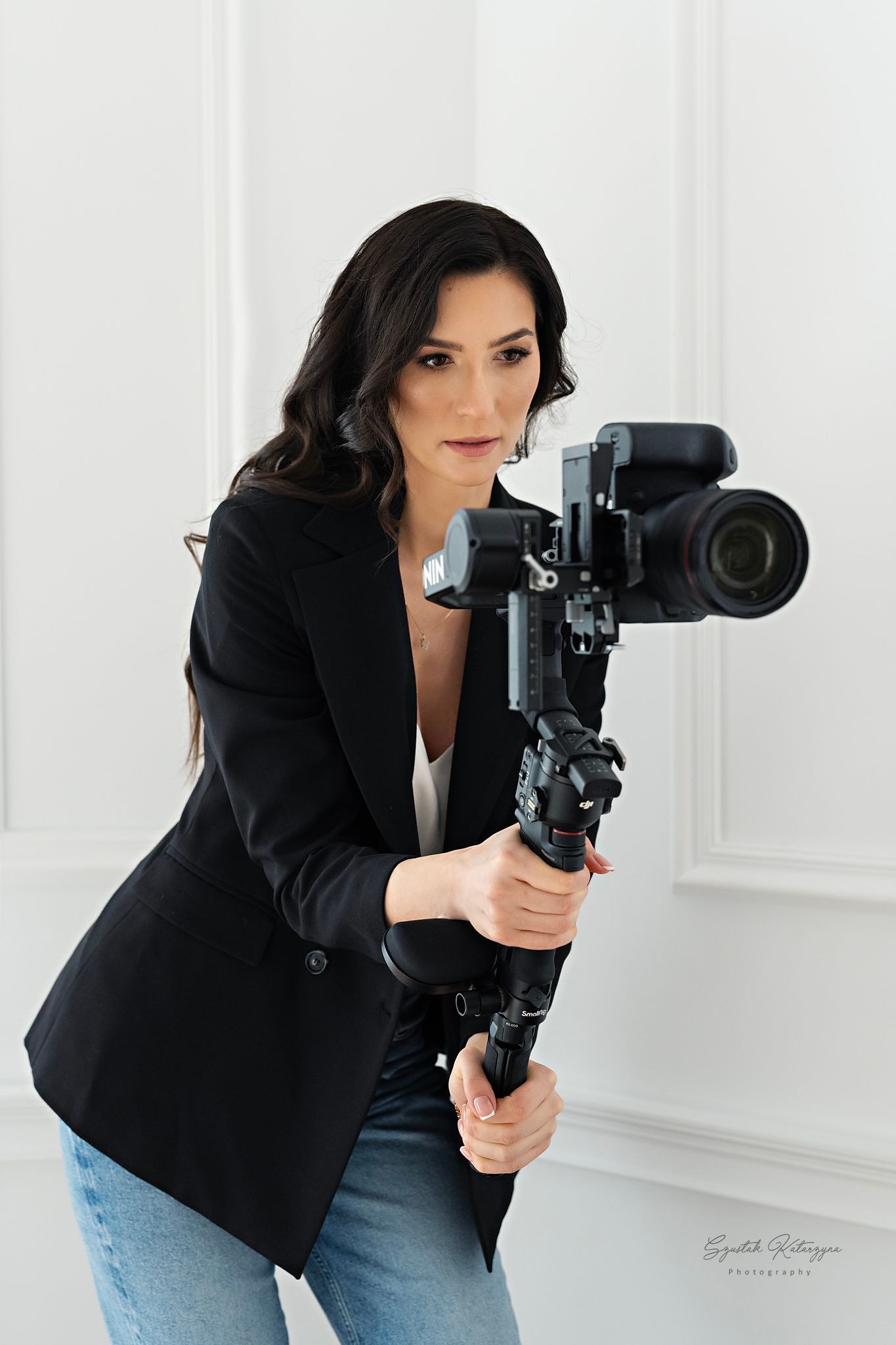 Kobieta w czarnym żakiecie trzyma stabilizator z aparatem, przygotowując się do nagrania. Jasne tło, profesjonalny sprzęt.