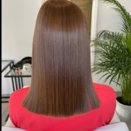 Proste, brązowe włosy po zabiegu prostowania, widok z tyłu. W tle lustro, roślina doniczkowa i stolik z filiżanką w salonie.