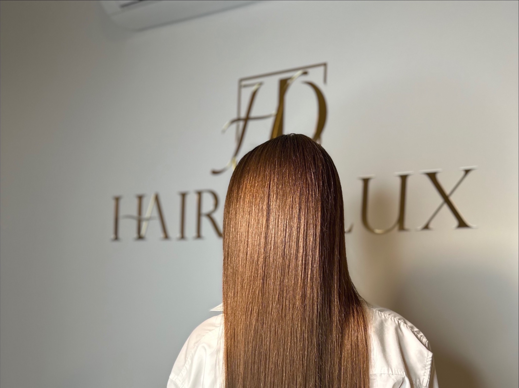 Proste, lśniące, brązowe włosy zakrywające twarz modelki, na tle ściany z logo 'HAIR LUX'. Efekt po zabiegu fryzjerskim.