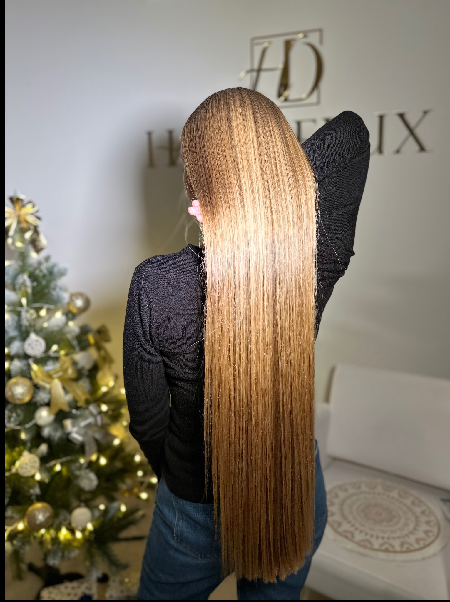 Długie, proste, blond włosy z delikatnymi refleksami, widoczne od tyłu, z choinką i logo salonu w tle. Efekt koloryzacji i pielęgnacji.