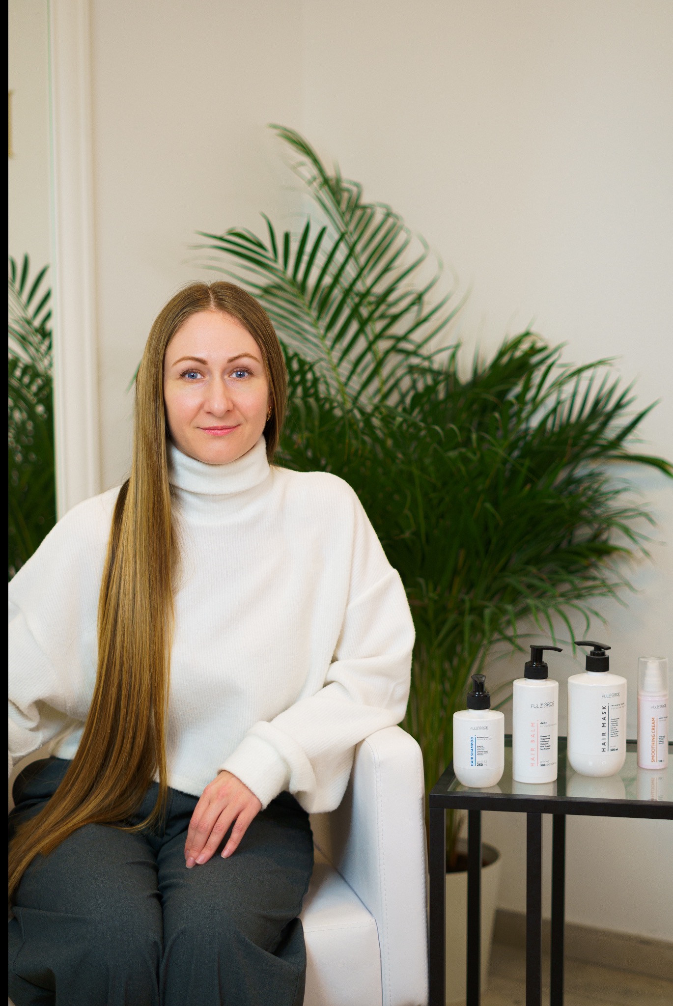 Kobieta z długimi, prostymi włosami siedzi na krześle w salonie w Gdańsku, obok niej kosmetyki do włosów marki Fuliforce na szklanym stoliku.