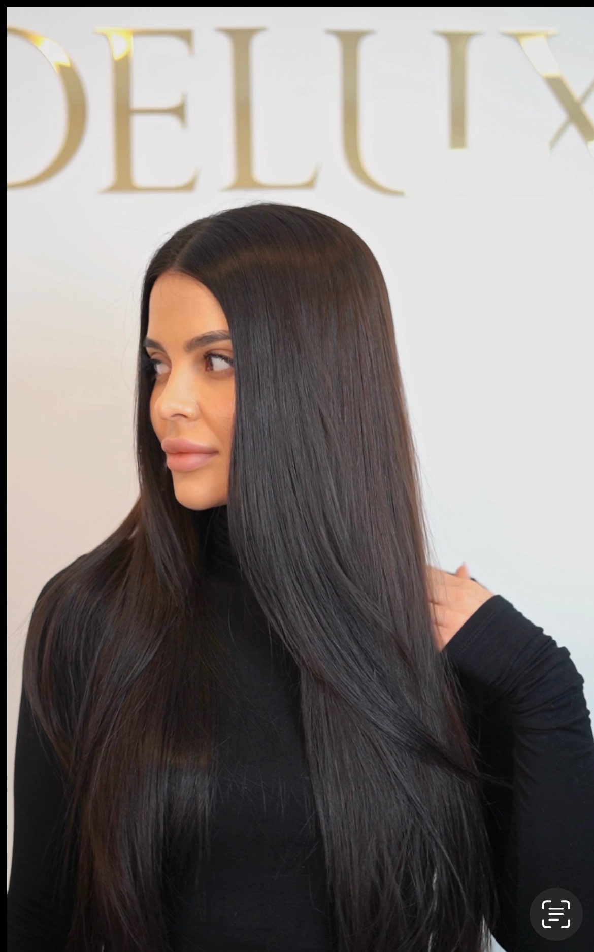 Kobieta z długimi, lśniącymi, czarnymi włosami, spoglądająca w lewo, na tle białej ściany z napisem 'DELUX' w złotym kolorze.