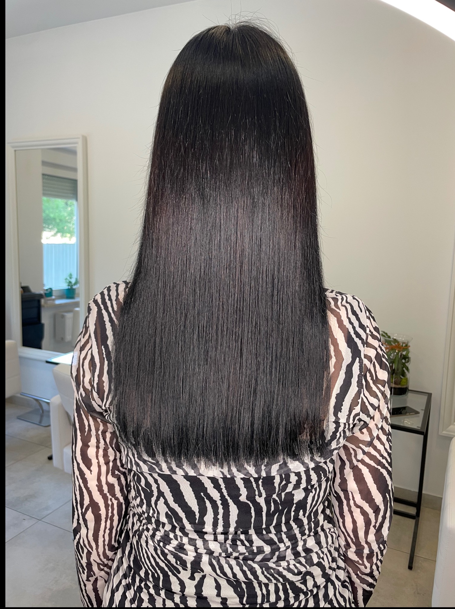 Długie, czarne, proste włosy na plecach kobiety w bluzce w zebrę. W tle jasne wnętrze salonu z lustrem.
