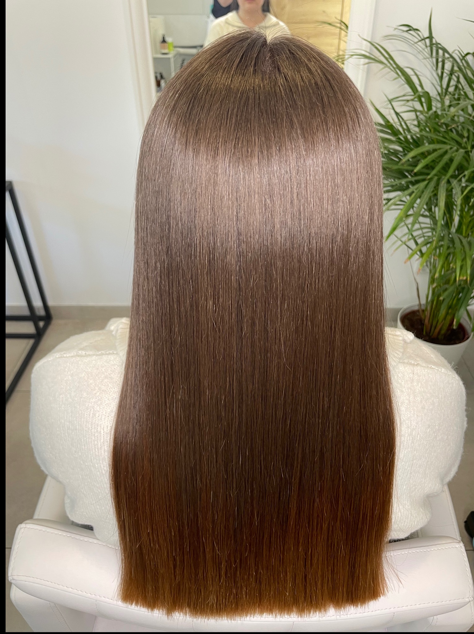 Gładkie, brązowe włosy po zabiegu prostowania, widok z tyłu, z delikatnym połyskiem, na tle rośliny doniczkowej i jasnego wnętrza salonu.