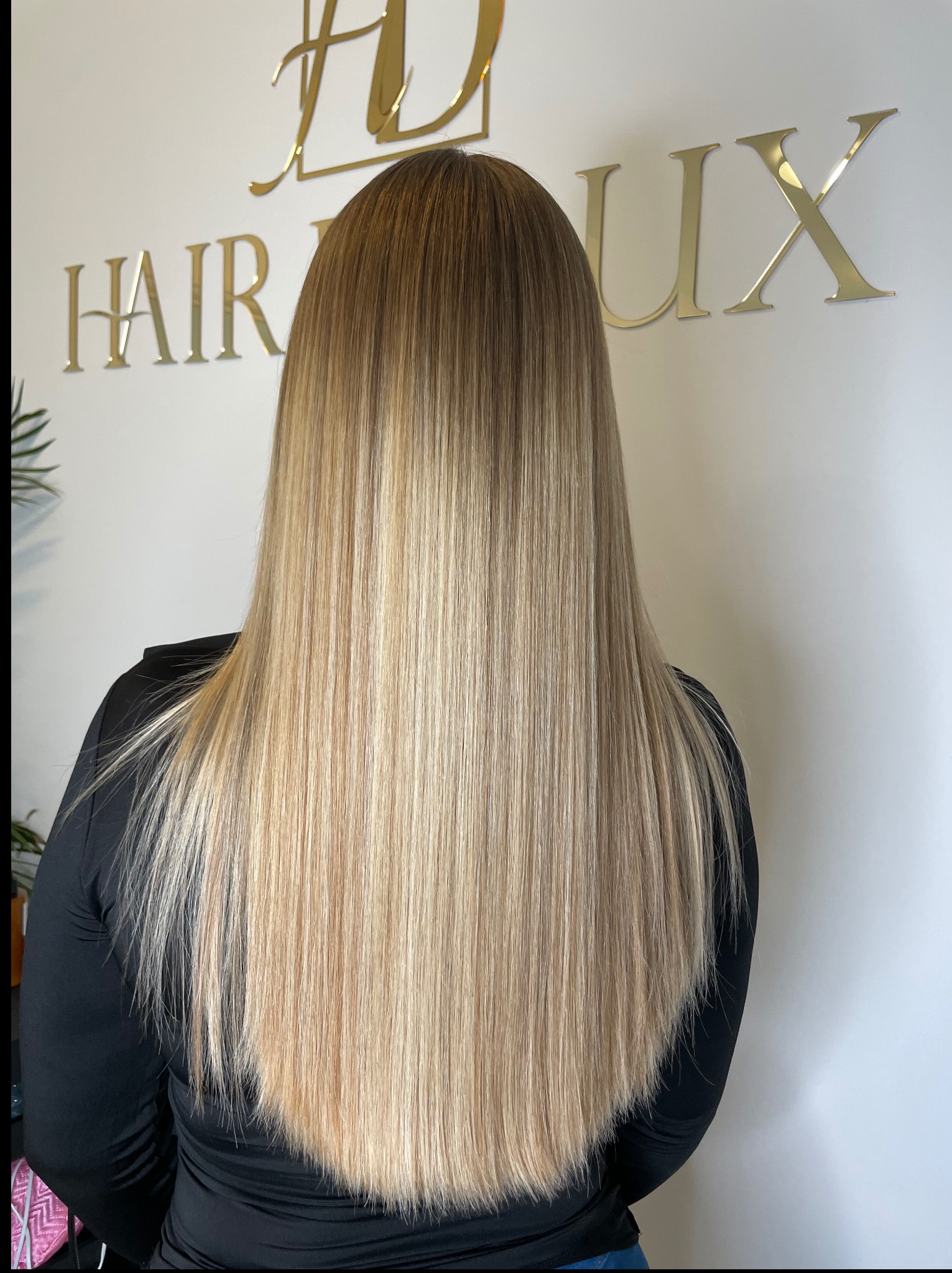 Długie, proste włosy z ombre w odcieniach blondu, widoczne od tyłu, na tle białej ściany z logo salonu fryzjerskiego Hair Lux.