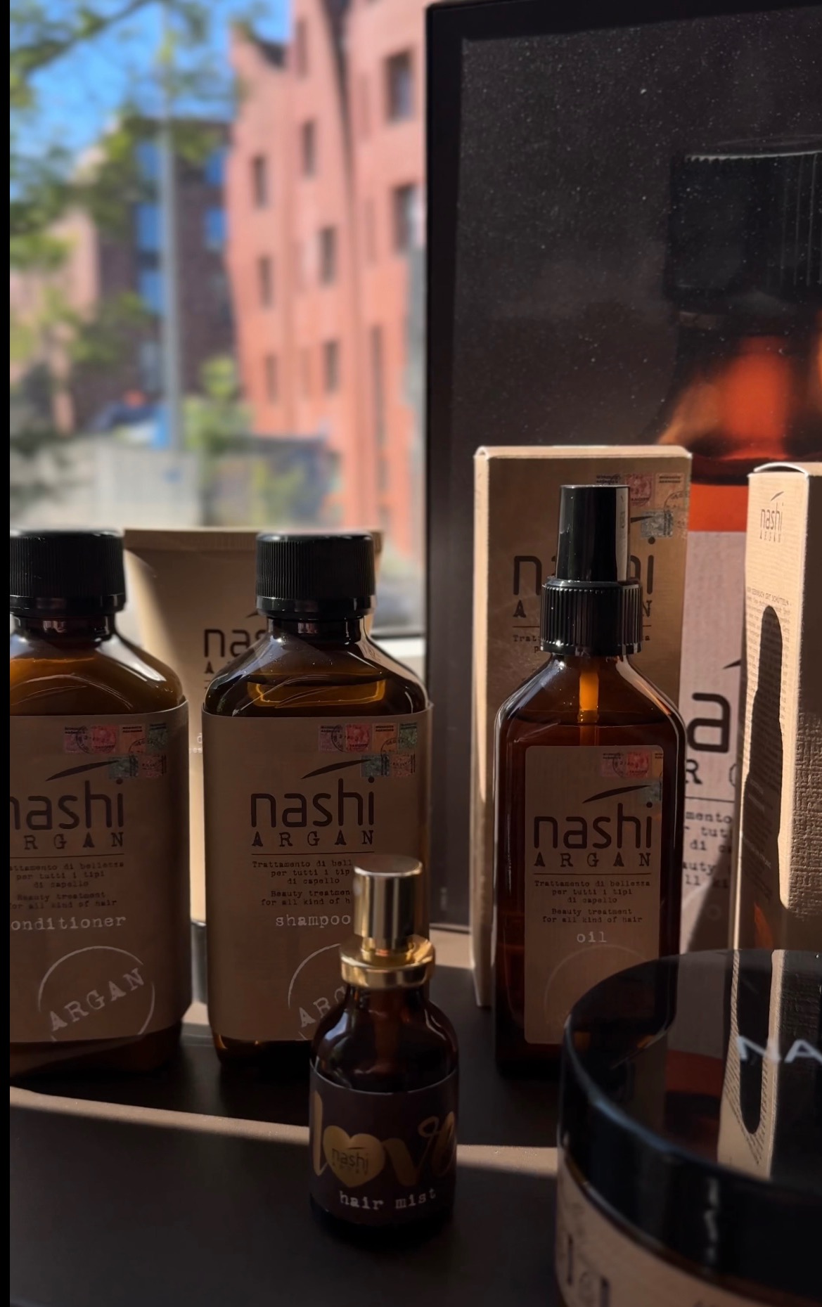 Zestaw kosmetyków Nashi Argan do pielęgnacji włosów: szampon, odżywka, olejek i hair mist. Produkty w brązowych butelkach na tle okna z widokiem na budynek.