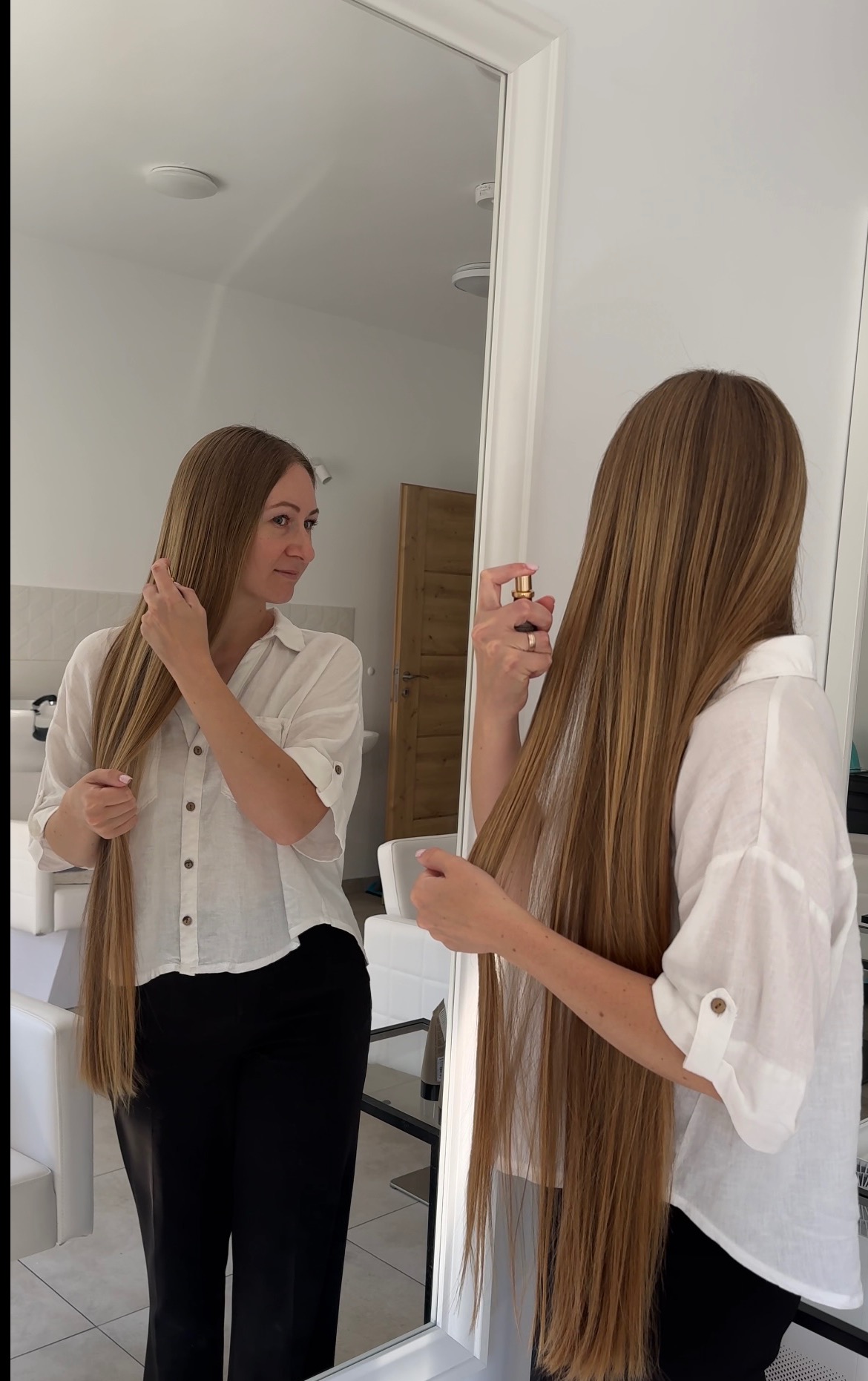 Kobieta z długimi, prostymi włosami spryskuje je preparatem, widoczna w lustrze w salonie. Jasne wnętrze, białe meble. Włosy sięgają poniżej pasa.