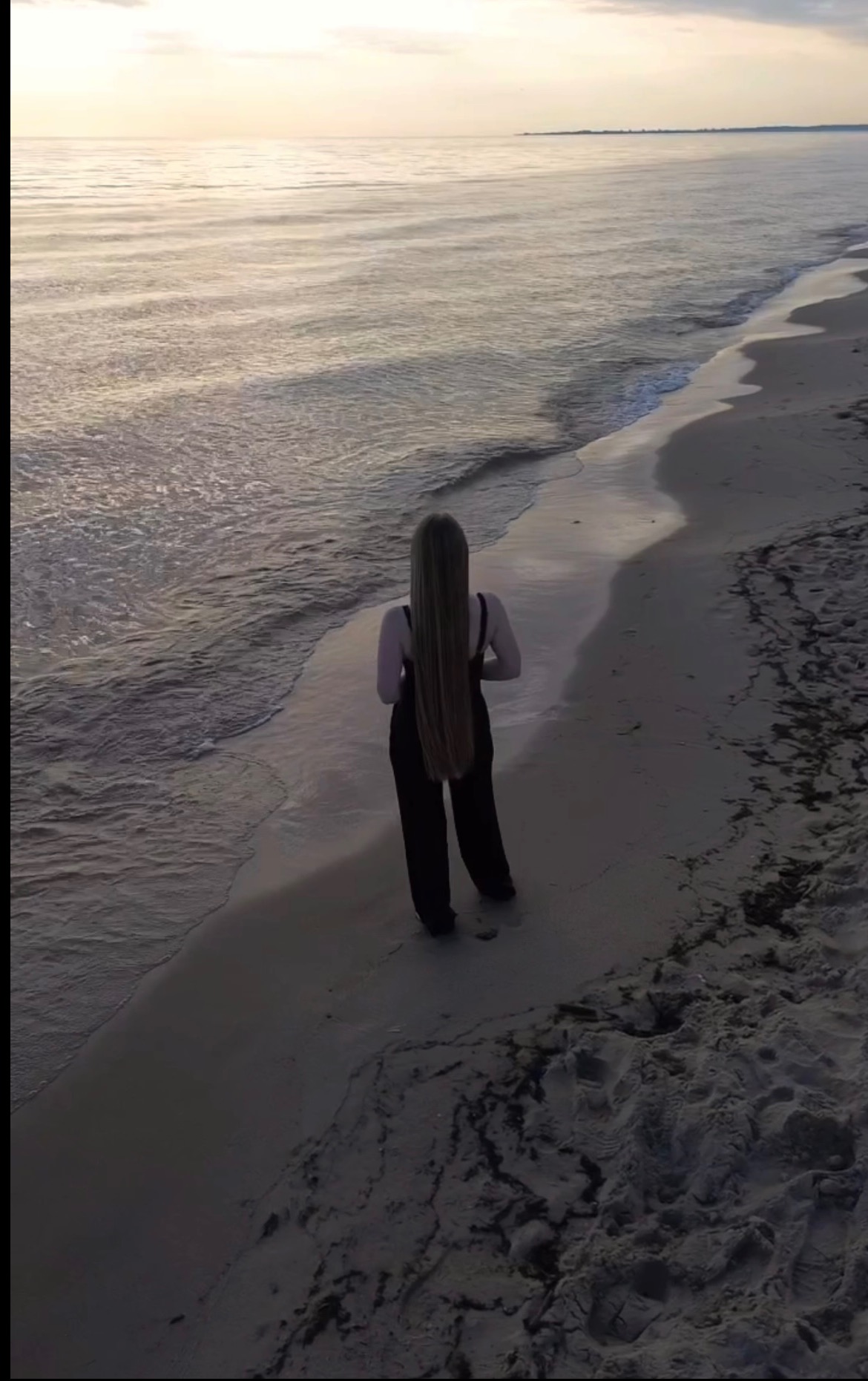 Kobieta w czarnym kombinezonie, tyłem, z długimi blond włosami na plaży o zmierzchu. Delikatne fale i piasek w odcieniach szarości i beżu.