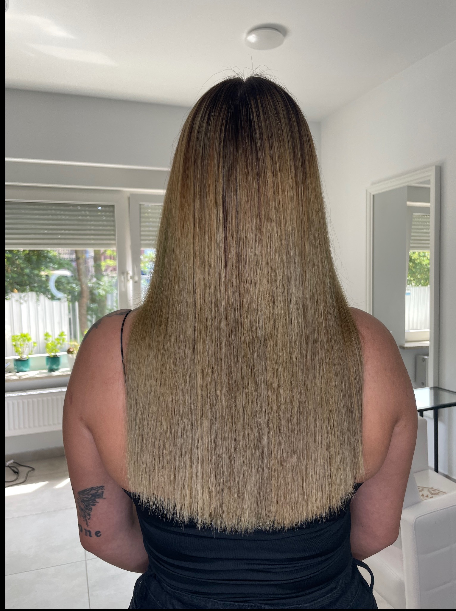 Długie, proste włosy po koloryzacji z ciemnymi odrostami, widok z tyłu, delikatne przejście kolorów, w tle jasne wnętrze z oknem i roślinami.