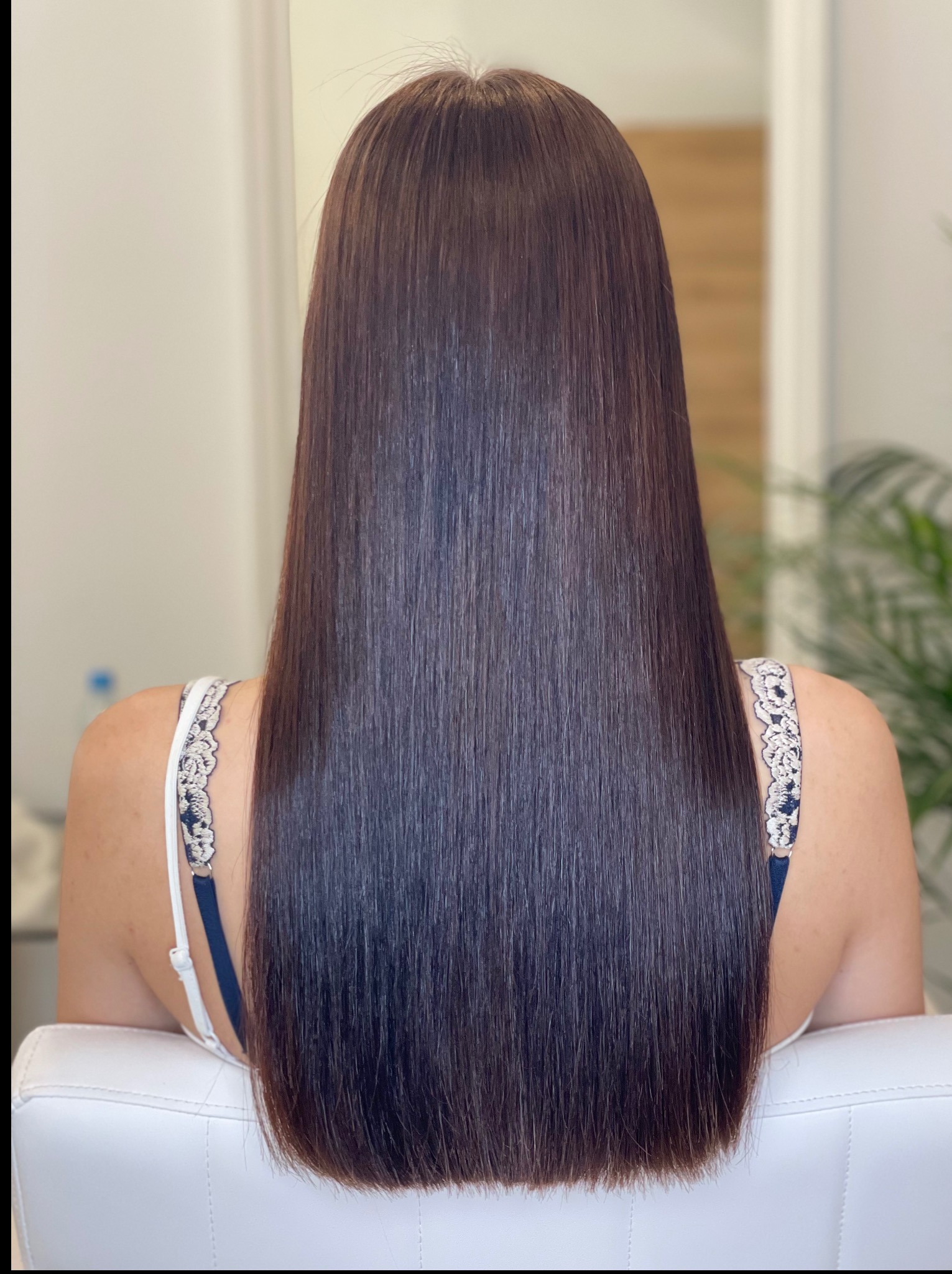 Proste, brązowe włosy do ramion, widok z tyłu na krześle. Włosy gładkie i lśniące, z delikatnym cieniowaniem na końcach. W tle widoczna zieleń rośliny.