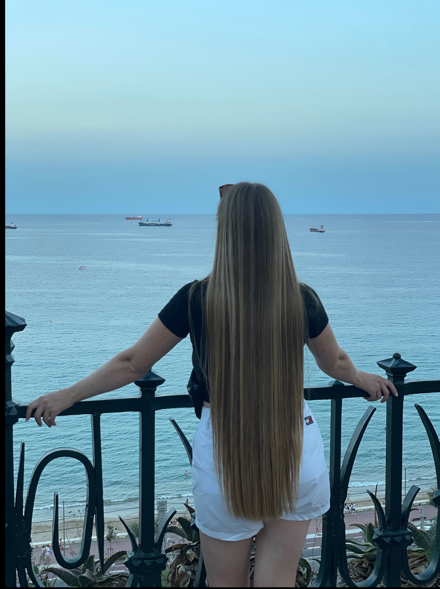 Kobieta z długimi, prostymi blond włosami, stojąca na balkonie z widokiem na morze, prezentująca efekt strzyżenia i pielęgnacji włosów.