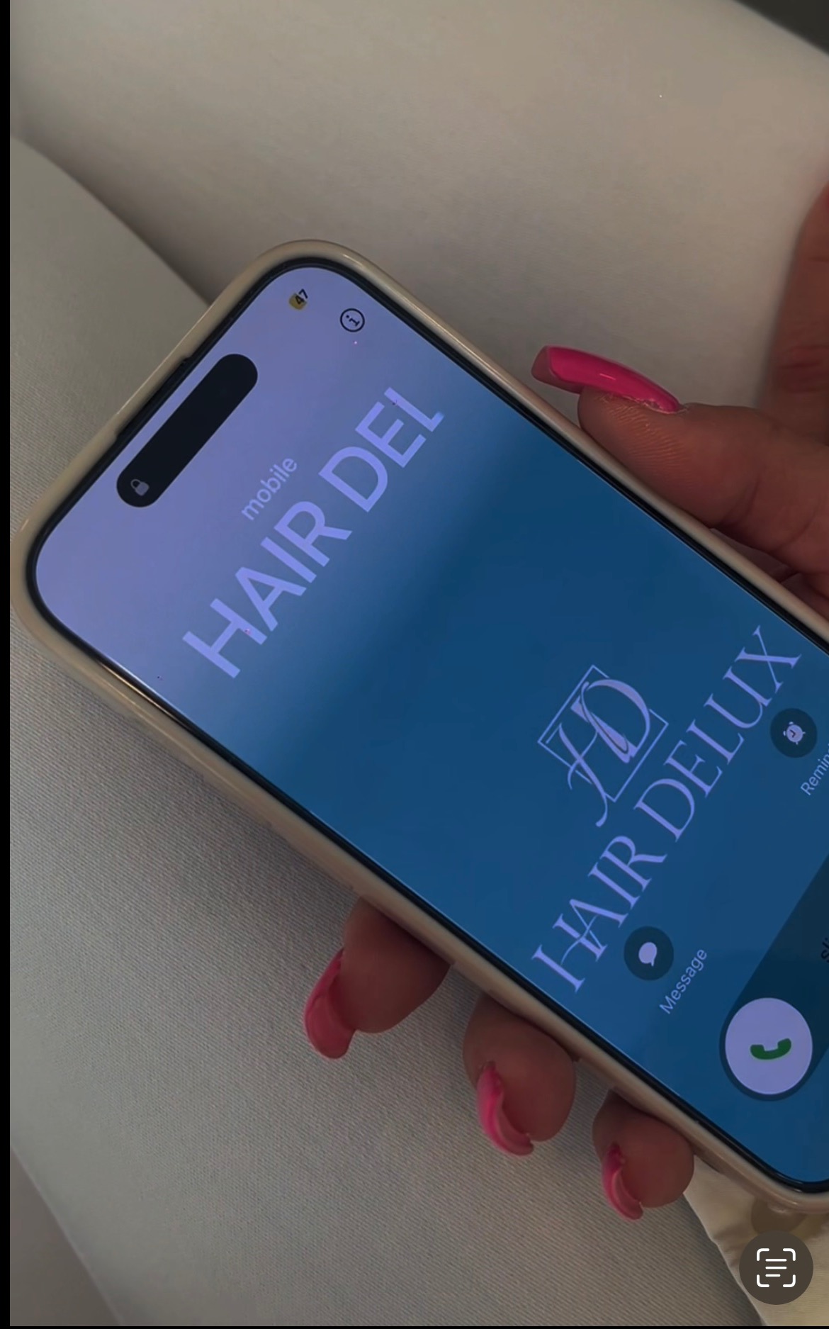 Smartfon z wyświetloną nazwą i logo 'Hair Delux' na ekranie połączenia przychodzącego, trzymany w dłoni z różowymi paznokciami.