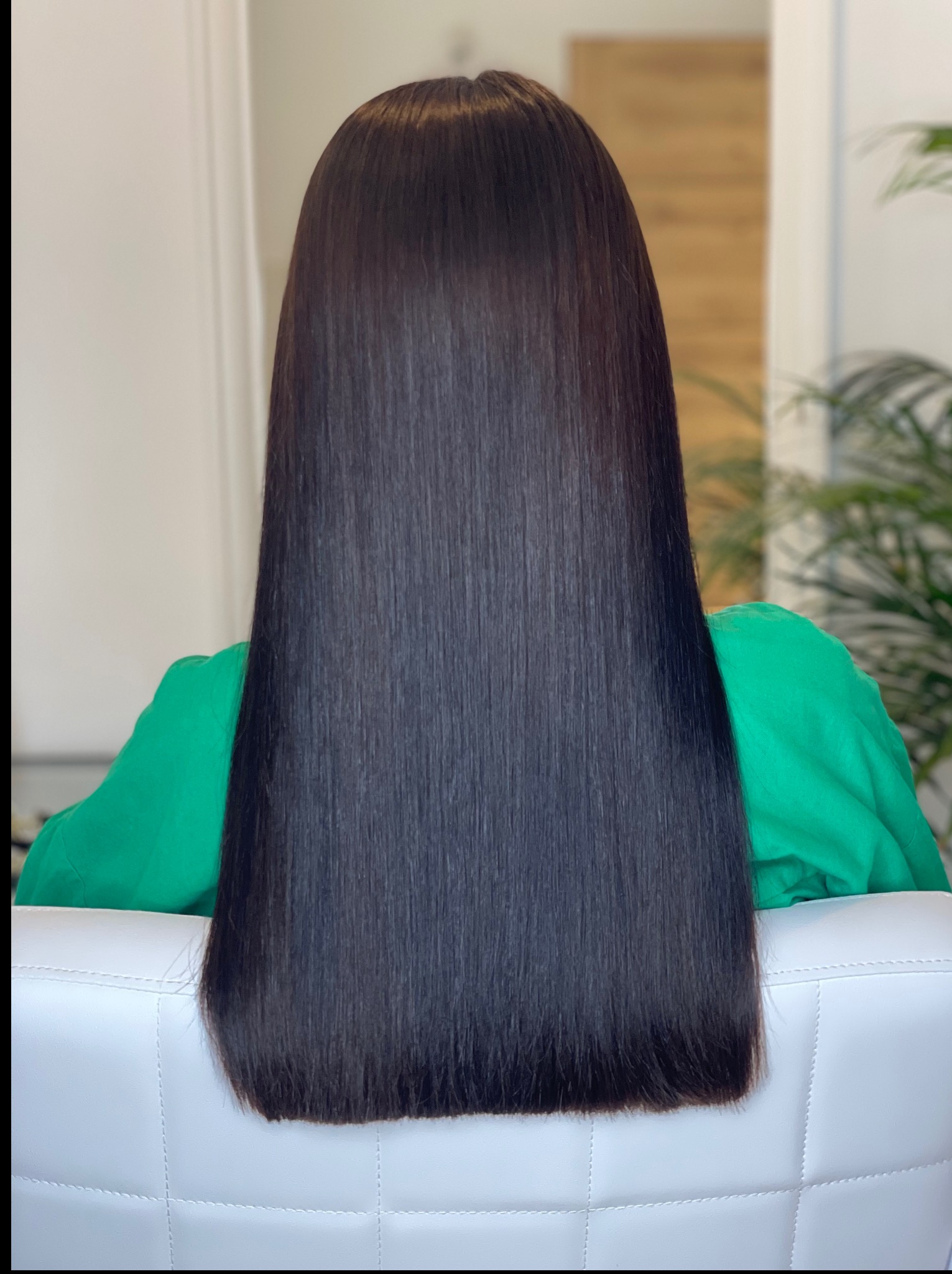 Długie, proste, ciemne włosy, widok z tyłu, po zabiegu prostowania. Włosy opadają na białe skórzane krzesło, klientka w zielonej bluzce, w tle roślina doniczkowa.
