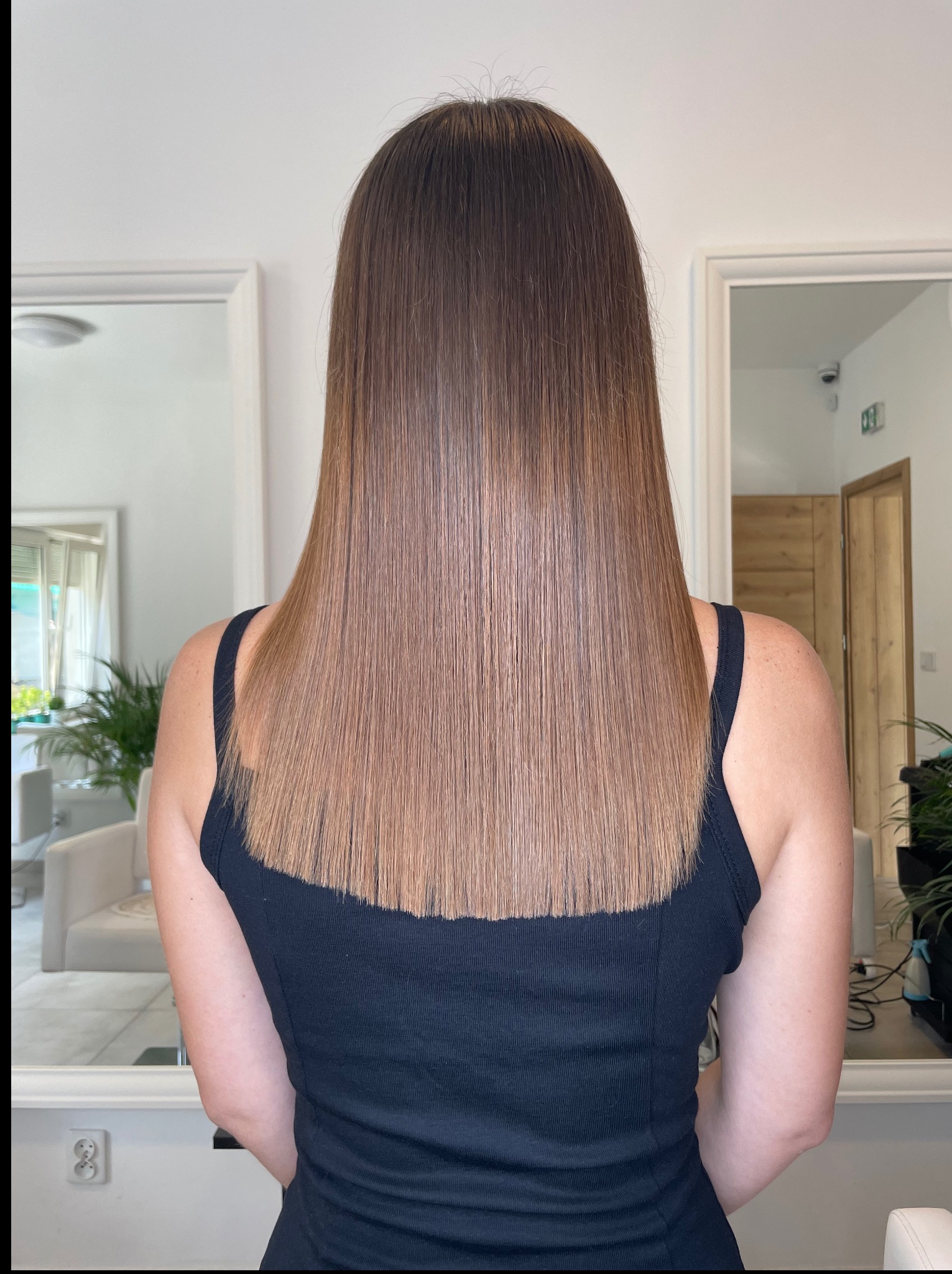 Proste, lśniące włosy w odcieniach brązu, świeżo po strzyżeniu. Widok z tyłu, w tle lustro i wnętrze salonu. Włosy sięgają do ramion, z delikatnym cieniowaniem.