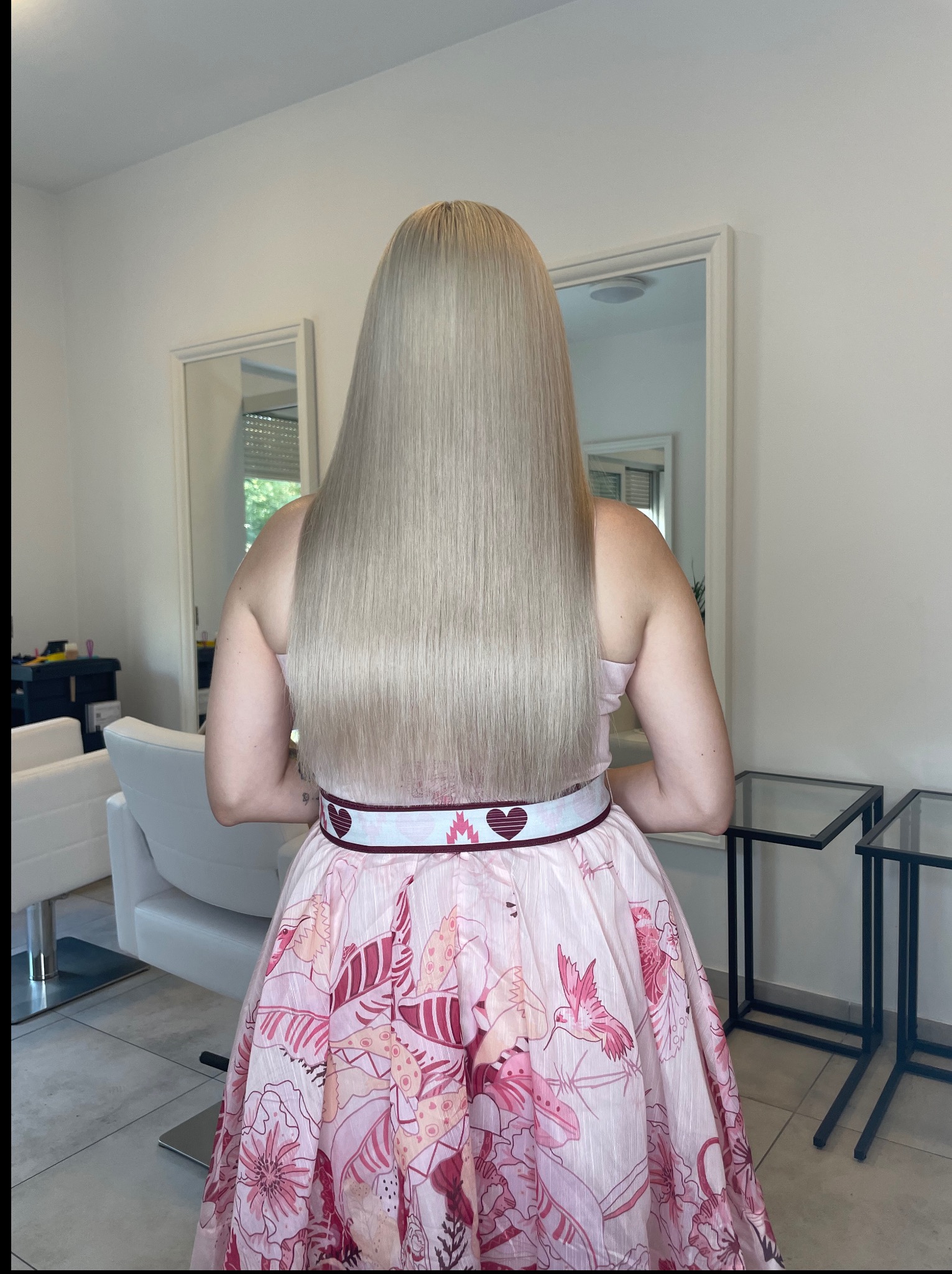 Blond włosy do pasa, wyprostowane i gładkie, widoczne z tyłu na tle salonu. Modelka w sukience w kwiaty, z paskiem w serca.