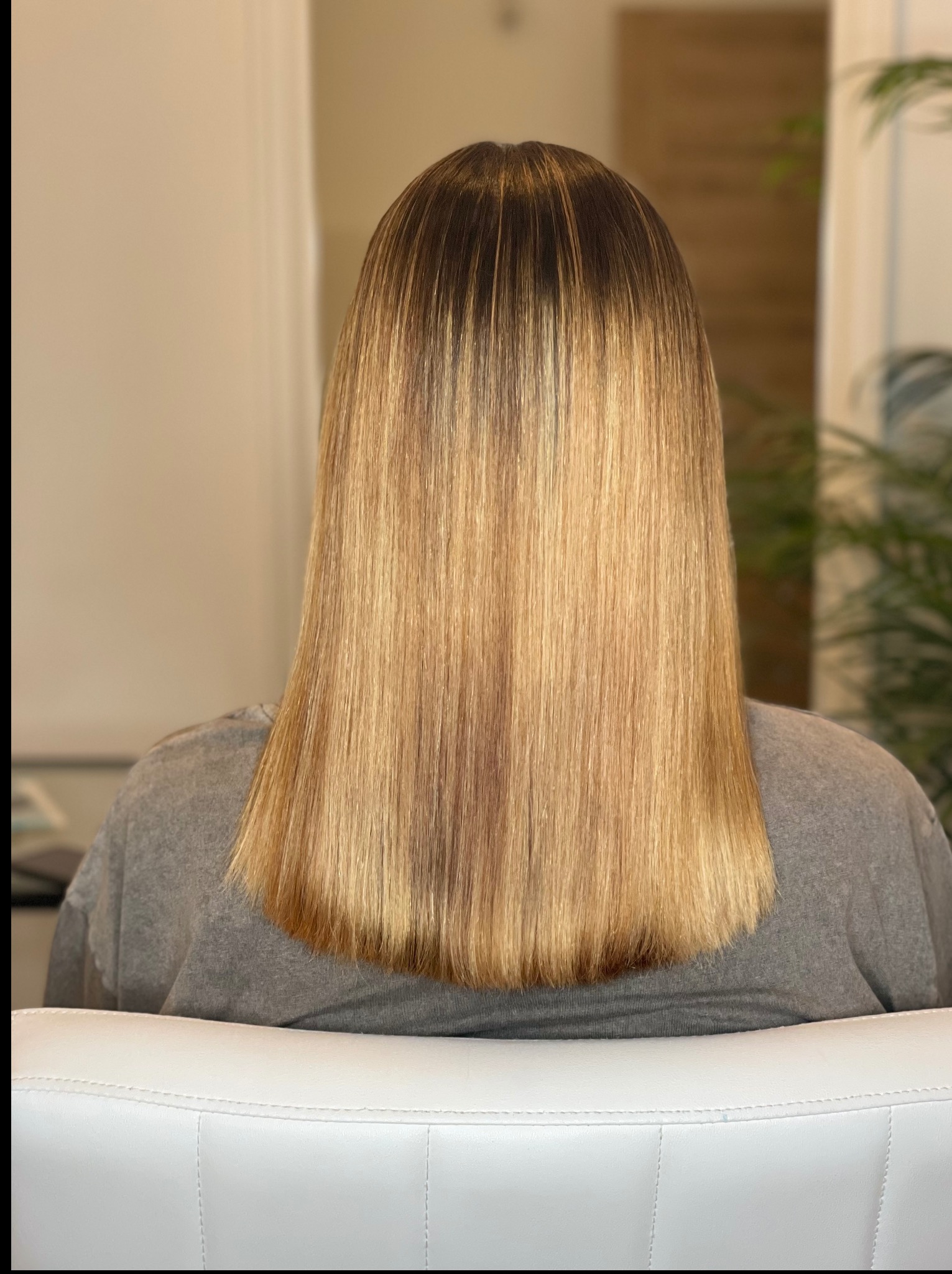 Proste, blond włosy z ciemniejszym odrostem, widok z tyłu na białym krześle. Włosy po zabiegu fryzjerskim, z połyskiem, na neutralnym tle.