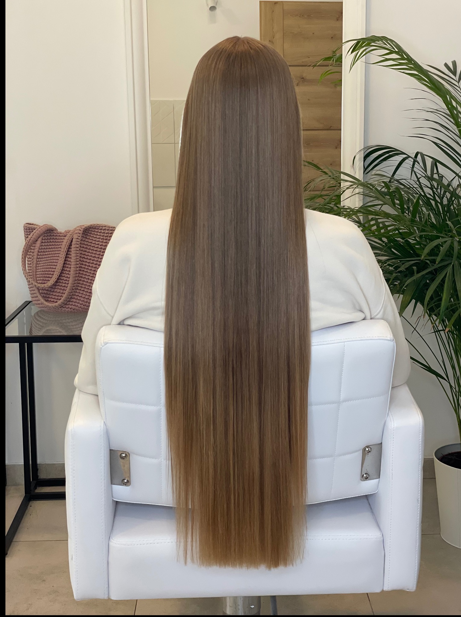 Długie, proste, brązowe włosy klientki po zabiegu koloryzacji, widok z tyłu. W tle jasne wnętrze salonu, białe krzesło i zielona roślina doniczkowa.