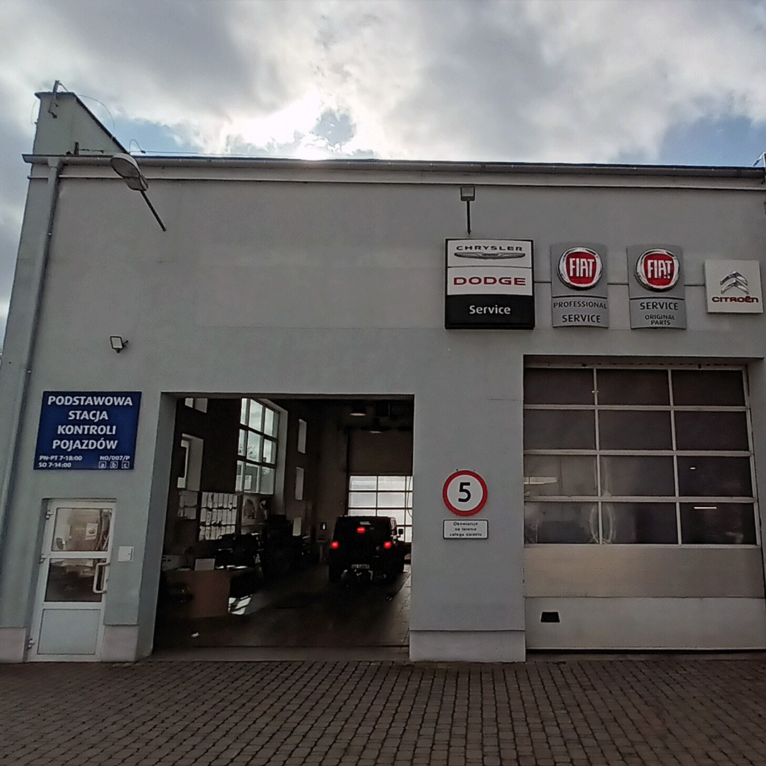 Fasada stacji kontroli pojazdów z logo marek Chrysler, Dodge, Fiat i Citroen. Wjazd do warsztatu, znak ograniczenia prędkości. Tablica informacyjna o godzinach pracy.