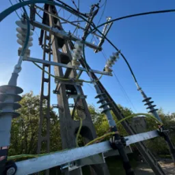 Słup energetyczny z widocznymi izolatorami i przewodami, sfotografowany pod kątem z dołu, na tle błękitnego nieba i zieleni drzew.