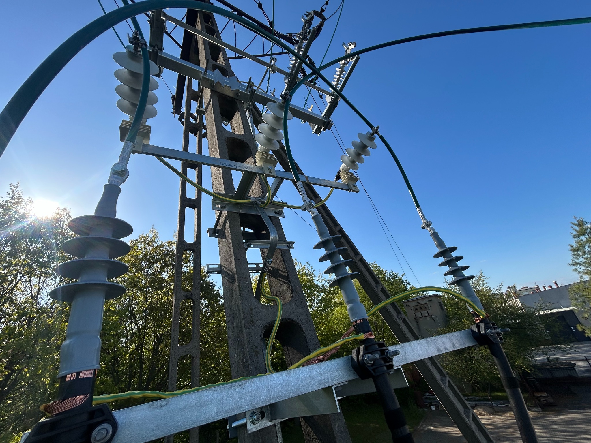 Słup energetyczny z widocznymi izolatorami i przewodami, sfotografowany pod kątem z dołu, na tle błękitnego nieba i zieleni drzew.