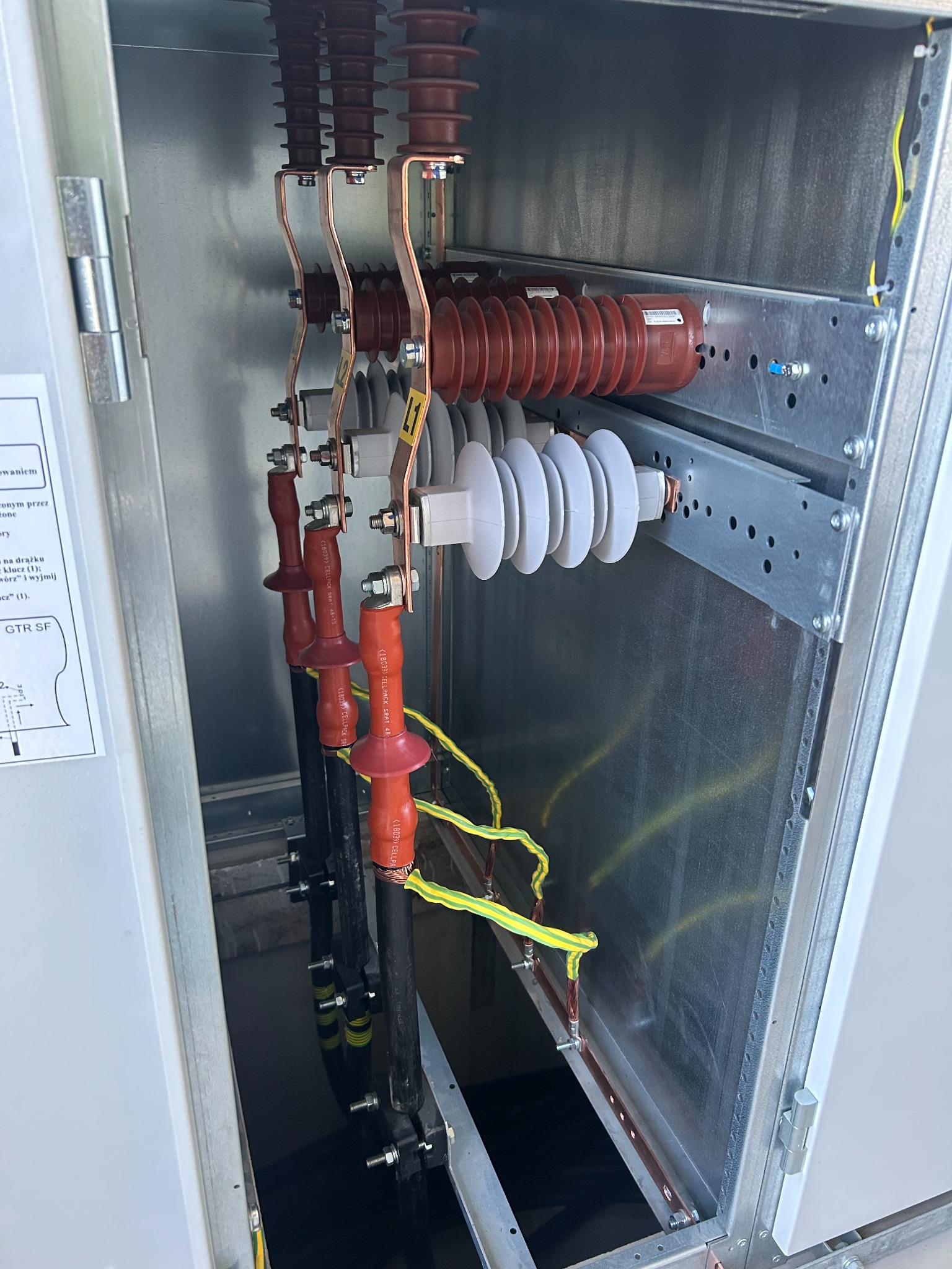 Rozdzielnica elektryczna z widocznymi elementami przyłączeniowymi, izolatorami i przewodami wewnątrz metalowej obudowy. Detal instalacji elektrycznej.