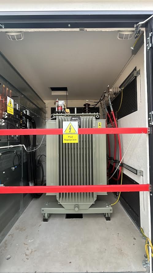 Wnętrze stacji transformatorowej z zainstalowanym transformatorem mocy, zabezpieczonym czerwoną barierą. Widoczne kable i tabliczki ostrzegawcze.