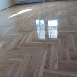 Parkiet ułożony w jodełkę, świeżo polakierowany, z wyraźnym odbiciem okna i światła słonecznego. Jasne drewno, błyszcząca powierzchnia.