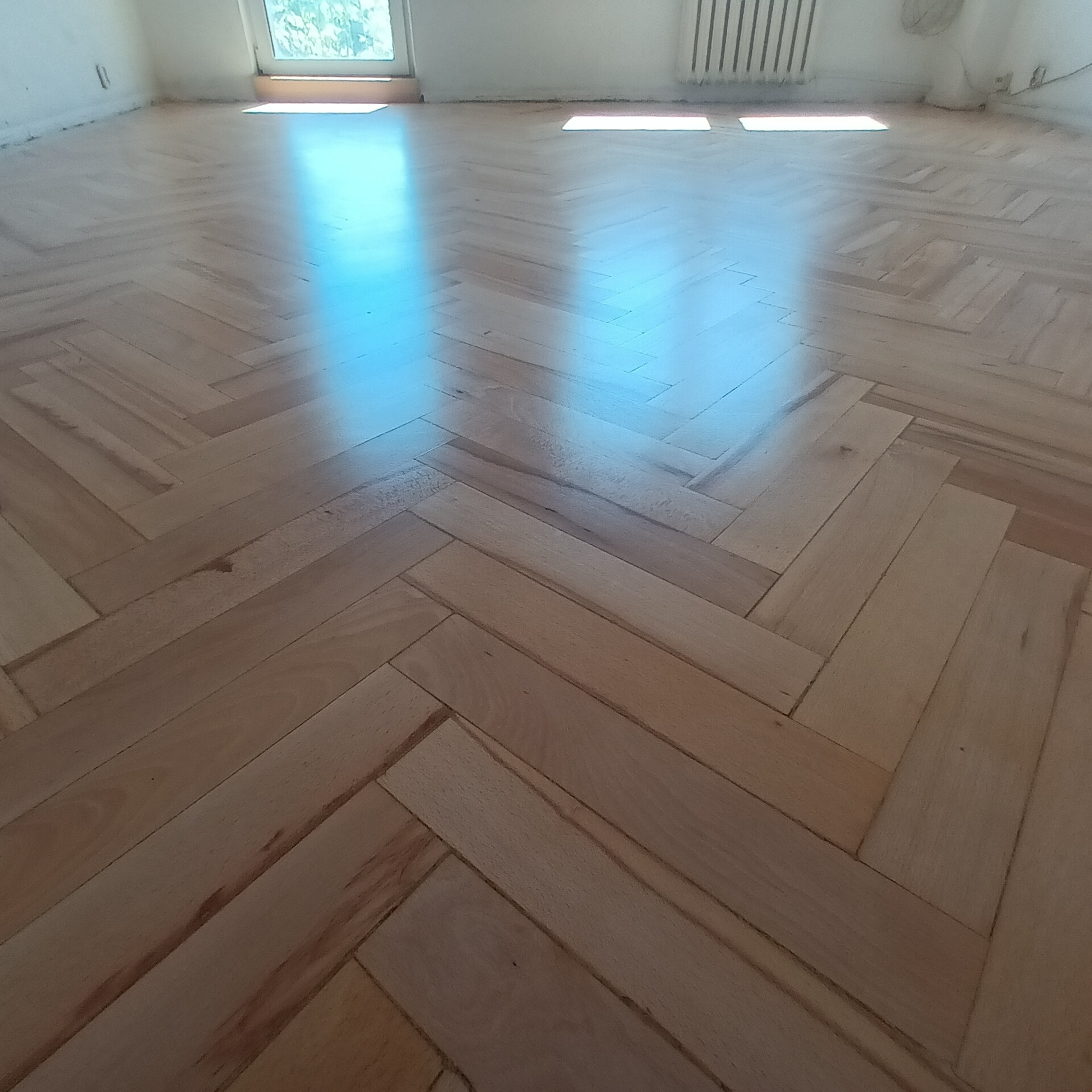 Jasny parkiet ułożony w jodełkę, odbijający światło okna. Widoczne detale naturalnego drewna i subtelne różnice w odcieniach poszczególnych klepek.