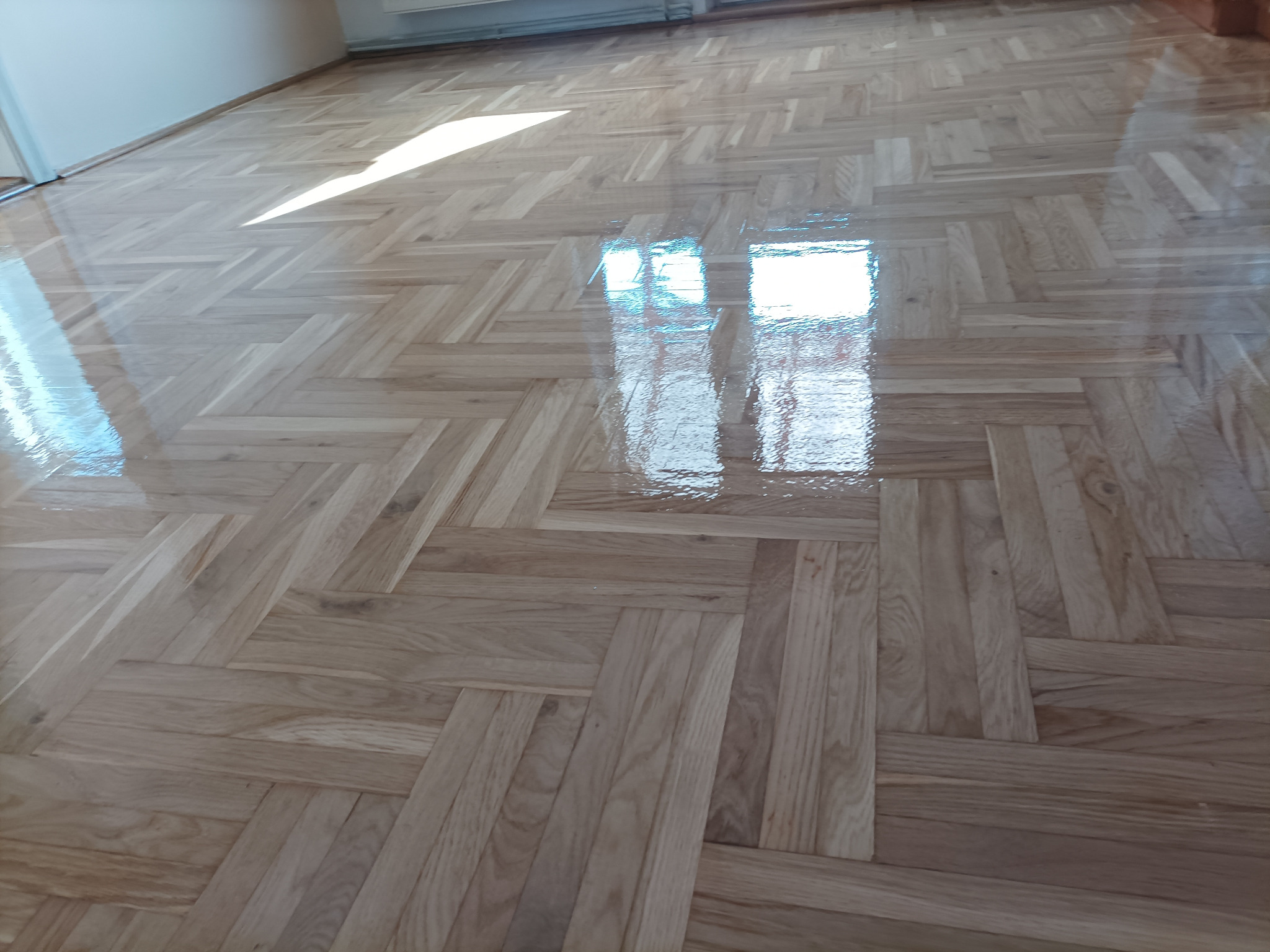 Parkiet ułożony w jodełkę, świeżo polakierowany, z wyraźnym odbiciem okna i światła słonecznego. Jasne drewno, błyszcząca powierzchnia.