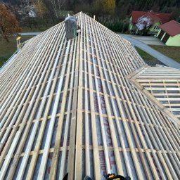 TOMHUS SPÓŁKA Z OGRANICZONĄ ODPOWIEDZIALNOŚCIĄ - Konstrukcja drewniana dachu z łatami, widok z góry na membranę i elementy mocujące. W tle zabudowa jednorodzinna i drzewa.
