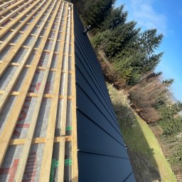 TOMHUS SPÓŁKA Z OGRANICZONĄ ODPOWIEDZIALNOŚCIĄ - Połowa dachu z czarnym blachy na rąbek, druga połowa w trakcie budowy z drewnianą konstrukcją i folią. W tle drzewa i błękitne niebo.