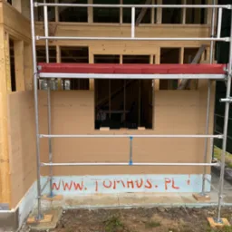 Konstrukcja domu w trakcie budowy, widoczne rusztowanie, drewniane elementy szkieletu i płyty Steico. Na fundamencie napis 'www.tomhus.pl'.