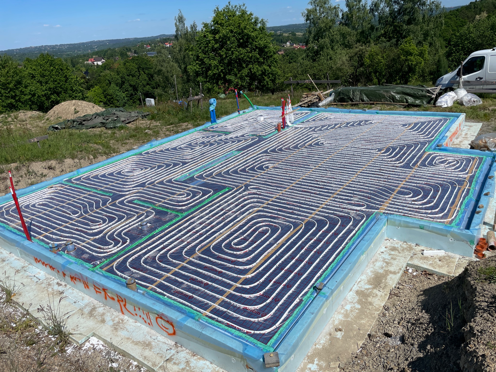 Fundament z izolacją i systemem ogrzewania podłogowego w domu w budowie. Widok z góry na ułożone rury grzewcze, zielone elementy mocujące i niebieską izolację.