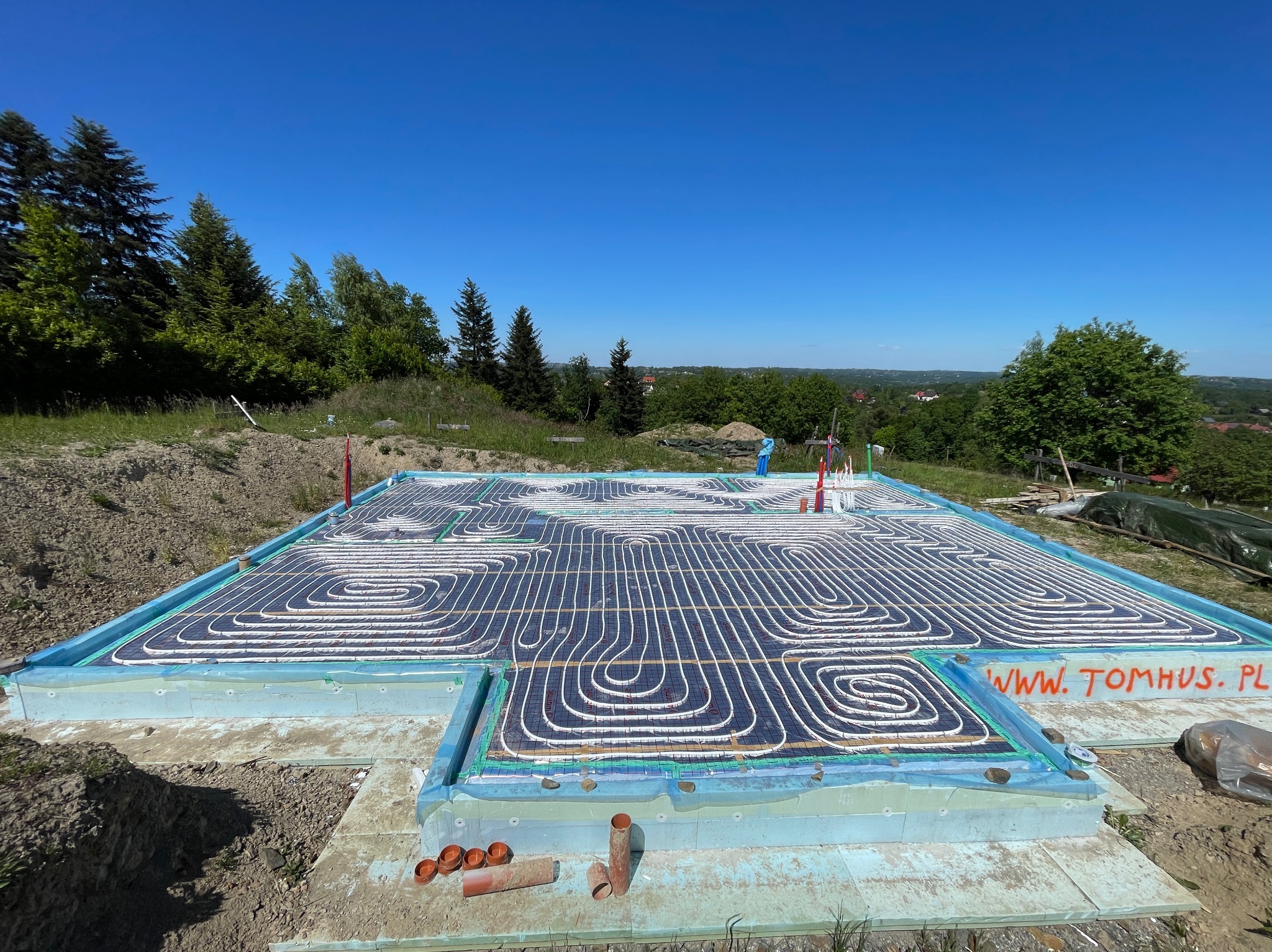 Instalacja ogrzewania podłogowego na fundamencie domu z izolacją, widok z góry. Na krawędzi widać napis 'www.tomhus.pl'.