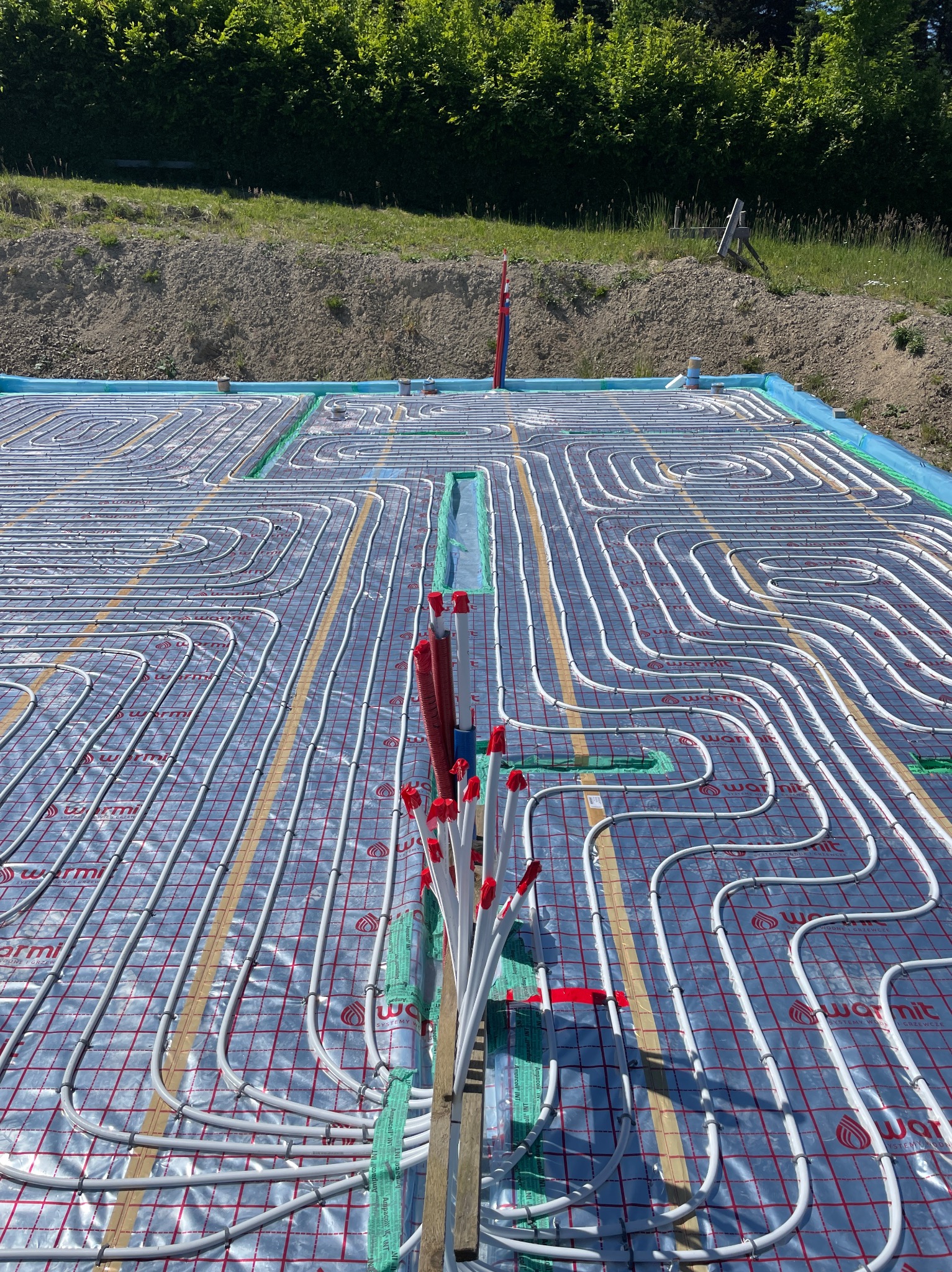 Instalacja ogrzewania podłogowego w domu szkieletowym/modułowym. Białe rurki ułożone na srebrnej macie z czerwoną siatką. Widoczne elementy mocujące i izolacja.