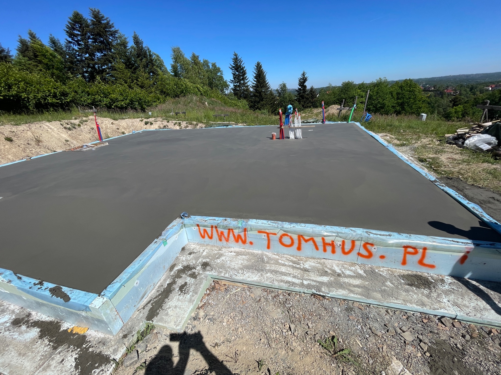 Wylana płyta fundamentowa pod budowę domu z widocznym adresem strony internetowej. W tle zieleń i błękitne niebo.