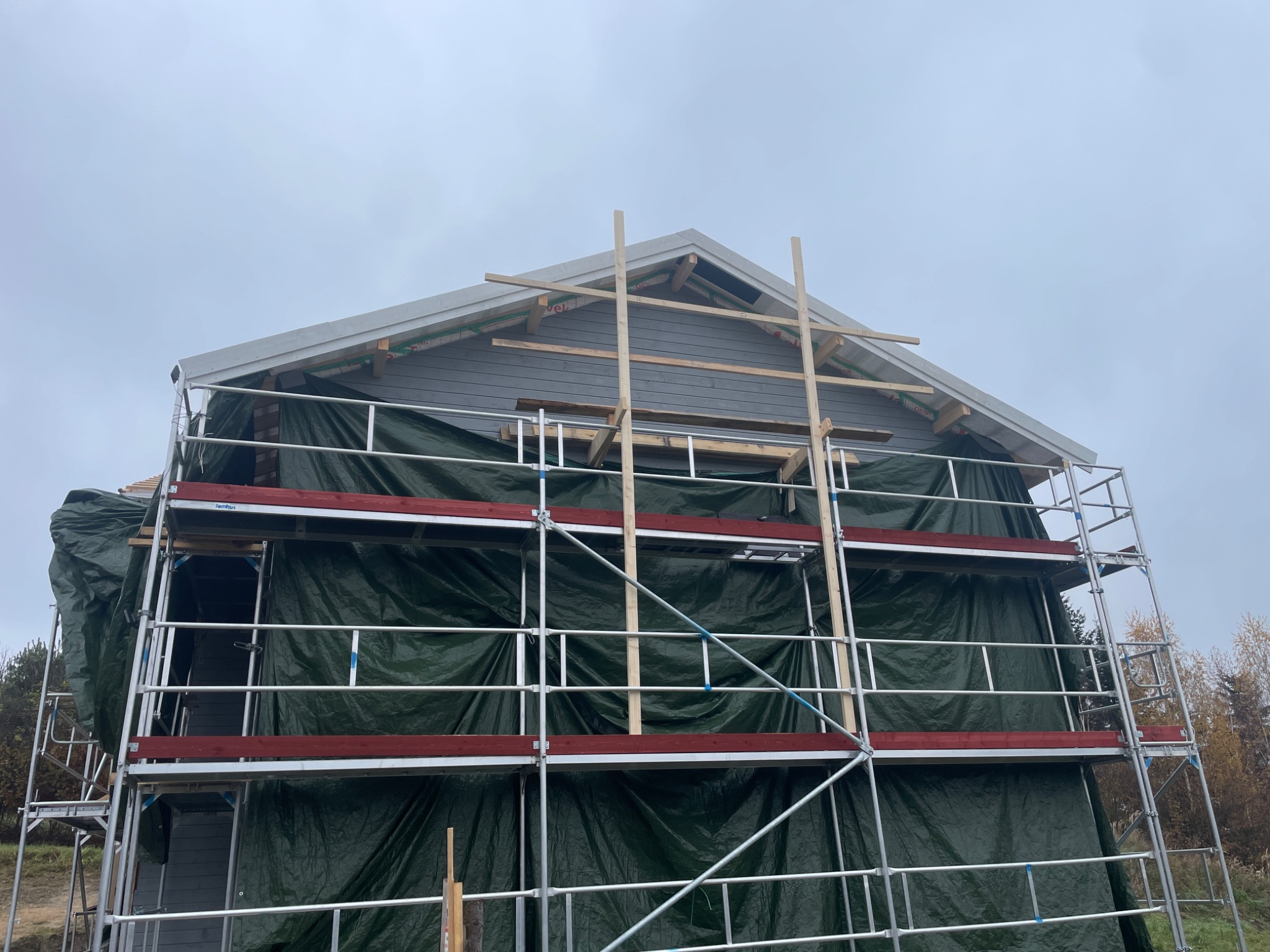 Budowa domu w trakcie: szkielet konstrukcji, rusztowania z zieloną plandeką, drewniane elementy więźby dachowej. Widok z dołu na pochmurne niebo.