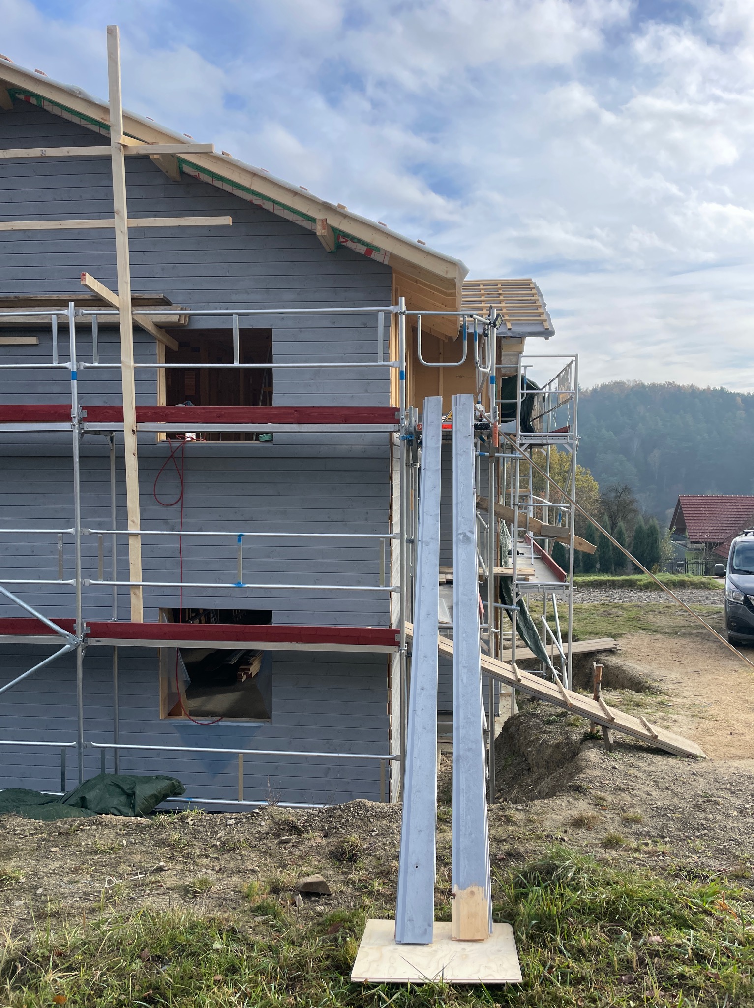 Szary dom w budowie z drewnianą konstrukcją dachu, otoczony rusztowaniami. Na pierwszym planie elementy elewacyjne i krajobraz z zalesionym wzgórzem w tle.